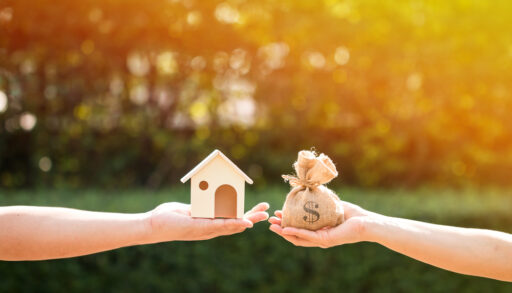 Two people holding a miniature wooden house and a small bag with a dollar sign on it.
