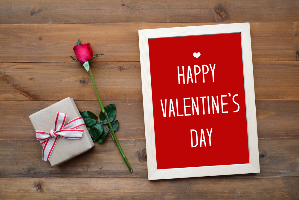 Red Valentine's Day sign on a wooden table with a wrapped present and a rose.
