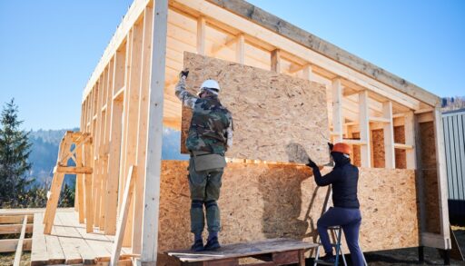 Two carpenters lifting a piece of plywood onto framing for a house.