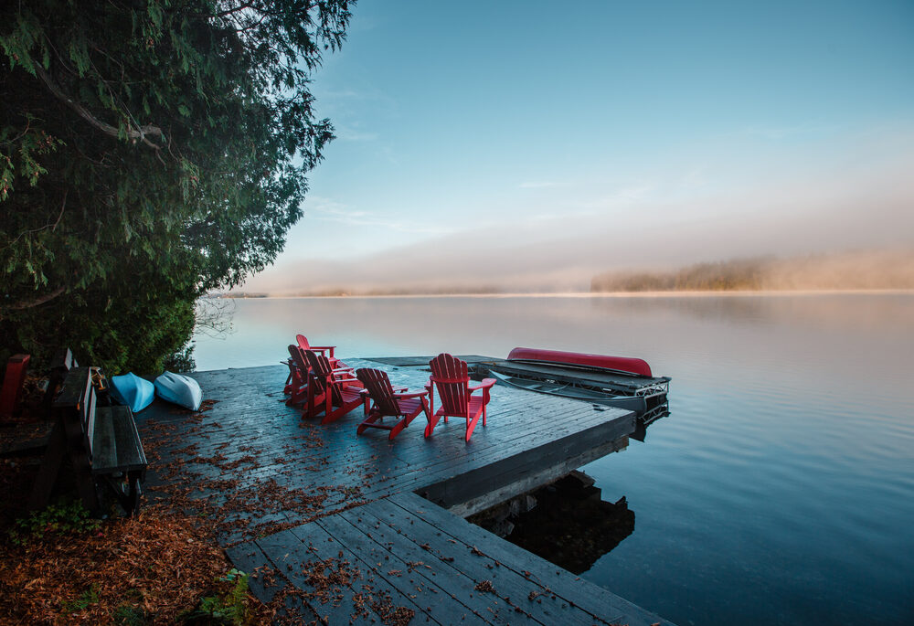 Red Muskoka chairs on a dock next to a still lake at sunset.