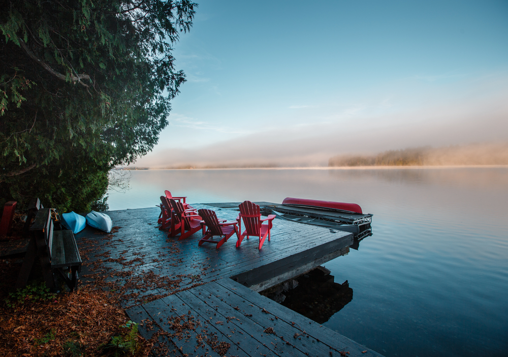 Red Muskoka chairs on a dock next to a still lake at sunset.