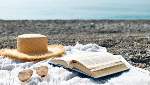 Hat, sunglasses and book laying open on a blanket next to water.