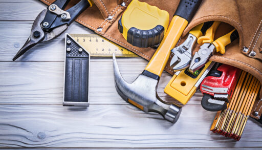 Tool belt with hammer, measuring tape and other tools laying on a table.