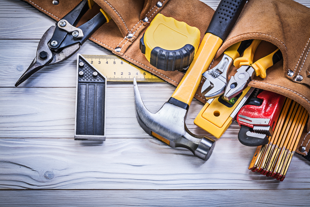 Tool belt with hammer, measuring tape and other tools laying on a table.