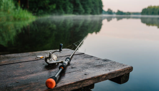 Fishing rod sitting on a wooden table overlooking a lake surrounded by trees.