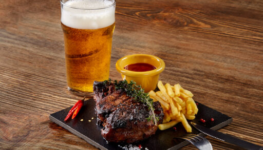 Glass of amber-coloured beer next to a plate with a steak and fries on it.