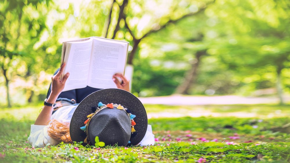 Woman wearing a grey hat reading a book in a field with trees.