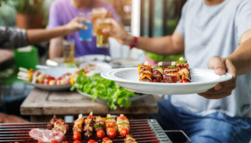 Man holding out a plate of food near a barbecue.