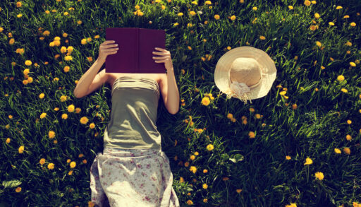 Girl laying in a field with yellow dandelions reading a book.