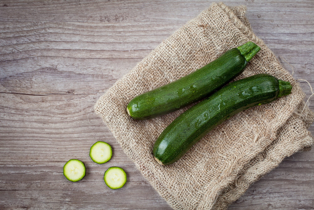 Overhead view of two green zucchini on a piece of burlap next to slices of zucchini.