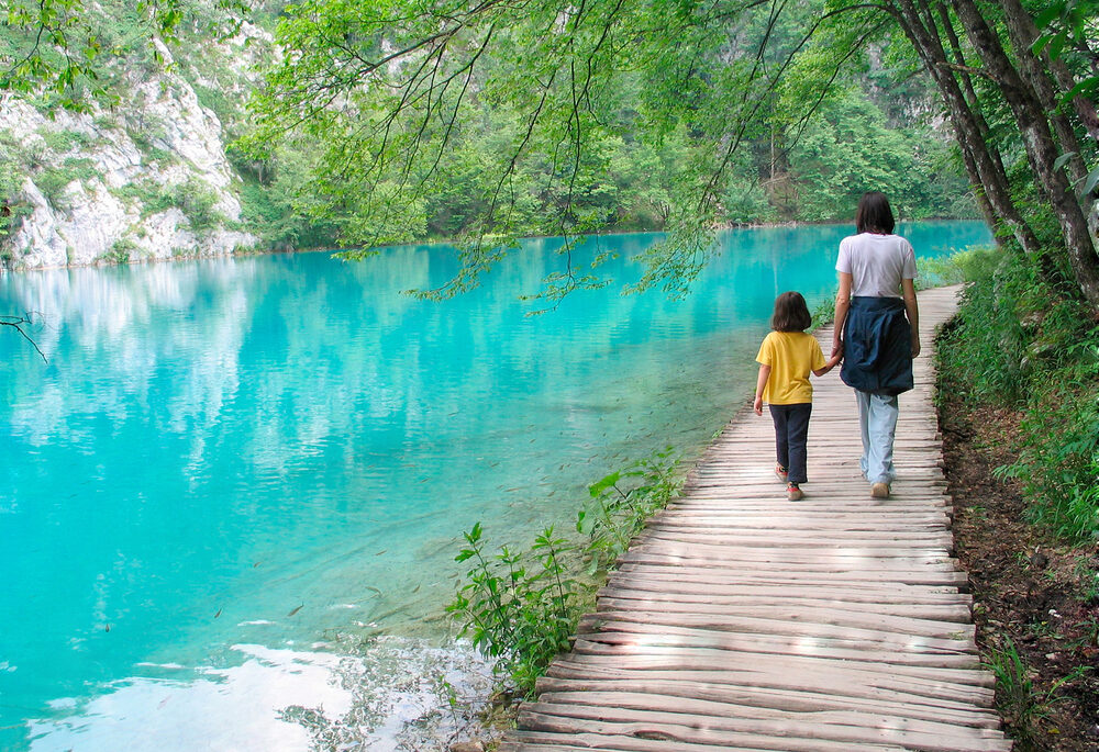 Woman and her child walk near a peaceful, turquoise-coloured lake surrounded by trees.