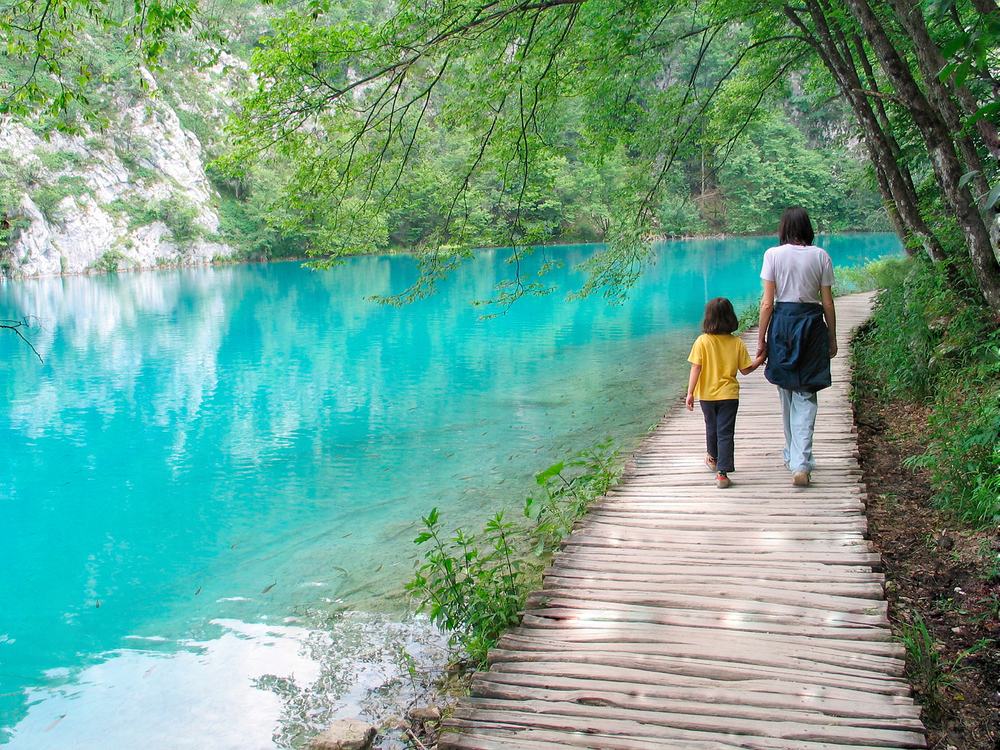 Woman and her child walk near a peaceful, turquoise-coloured lake surrounded by trees.