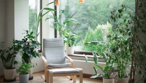 Grey armchair next to a big picture window surrounded by green plants.