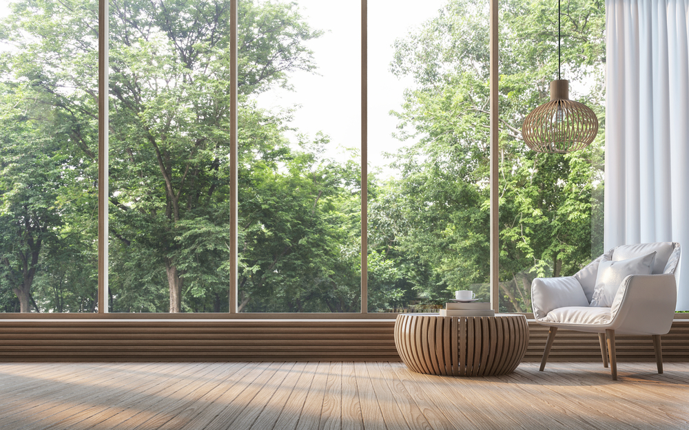 Modern living room with wooden table and grey chair in front of large, panoramic windows looking into a forest.