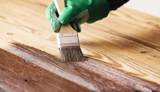 Close-up of a person wearing a green glove using a paintbrush to stain wood.