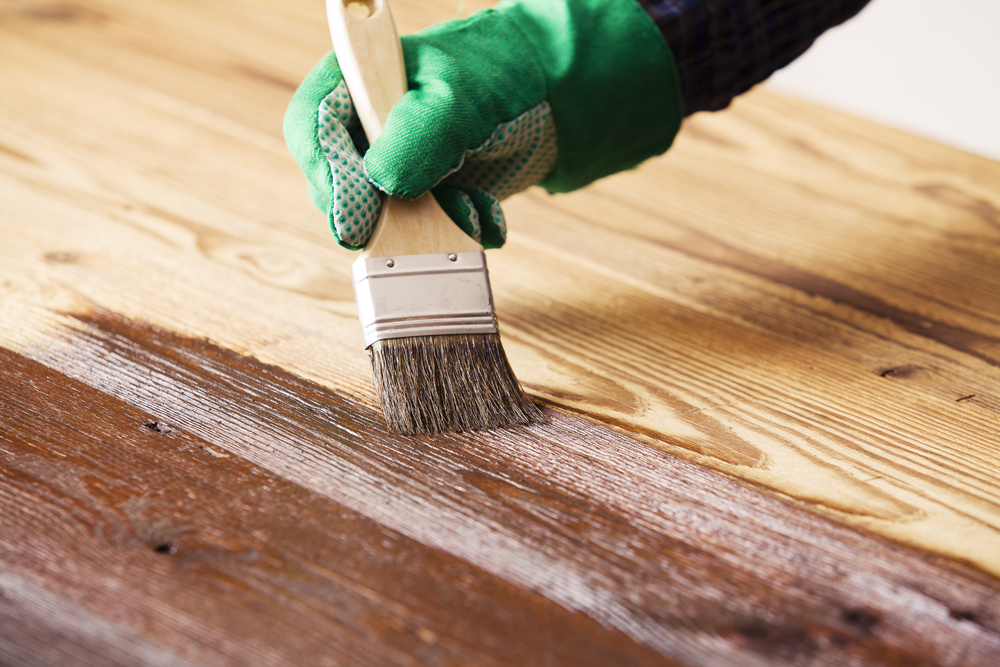 Close-up of a person wearing a green glove using a paintbrush to stain wood.