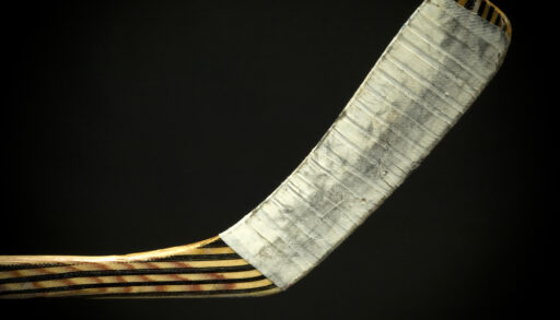 Close-up of the a wooden hockey stick blade with white tape and a black background.