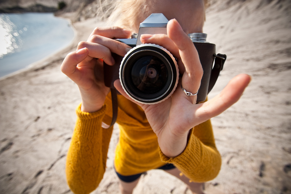 Woman in a yellow sweater holding an old camera and pointing it at the photographer.