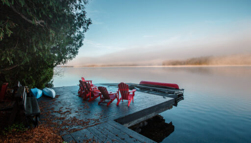 Red Muskoka chairs on a dock with a tree hanging over it.
