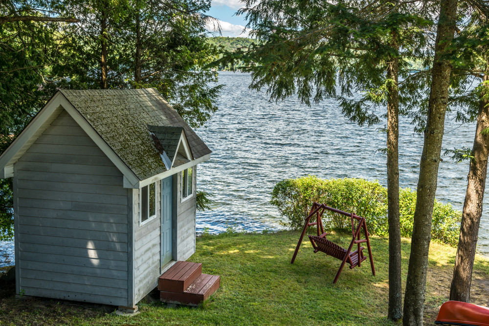 Small, light blue cottage bunkie next to a lake surrounded by trees.