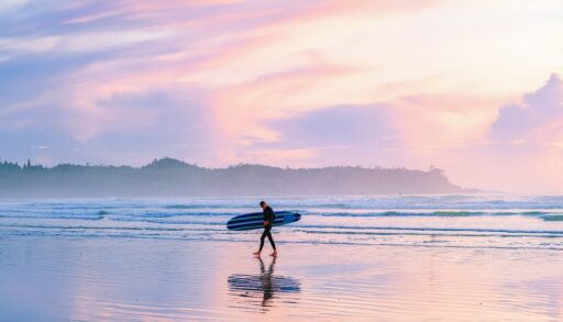 Person walking along Tofino Beach at sunset holding a surfboard.