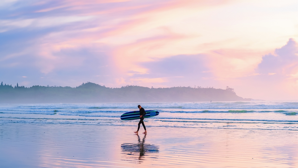 Person walking along Tofino Beach at sunset holding a surfboard.