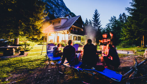 Group of people sitting around a campfire near a cottage in the evening.
