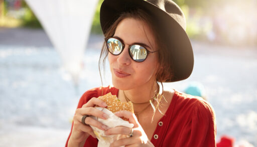 Woman in a hat and sunglasses holding and eating a hamburger.