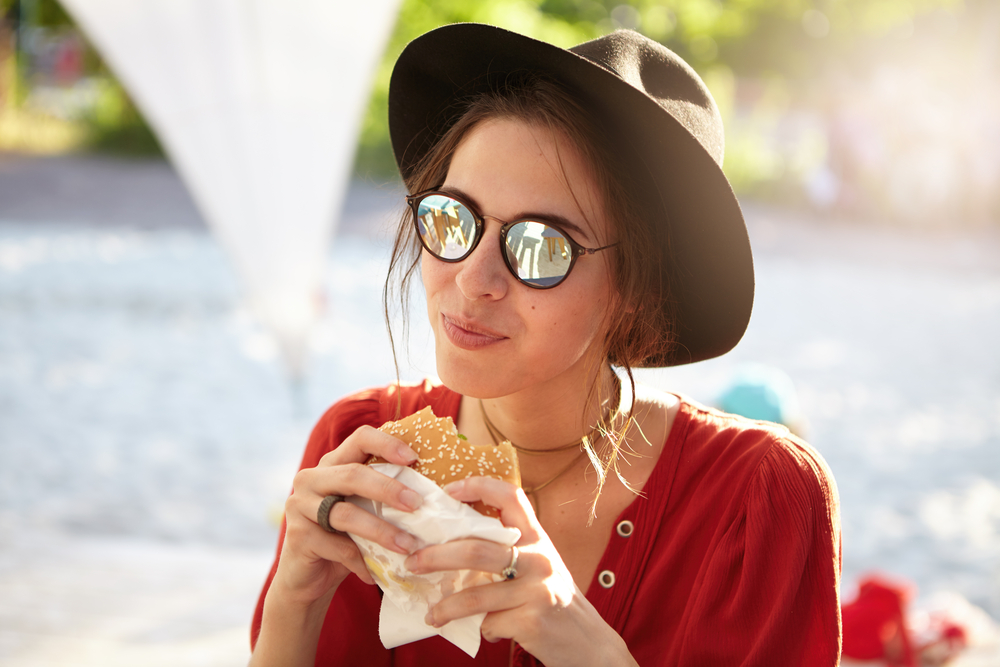 Woman in a hat and sunglasses holding and eating a hamburger.