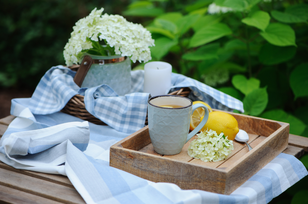 Wooden tray with a grey coffee mug on an outdoor table with a blue tablecloth and vase of flowers.