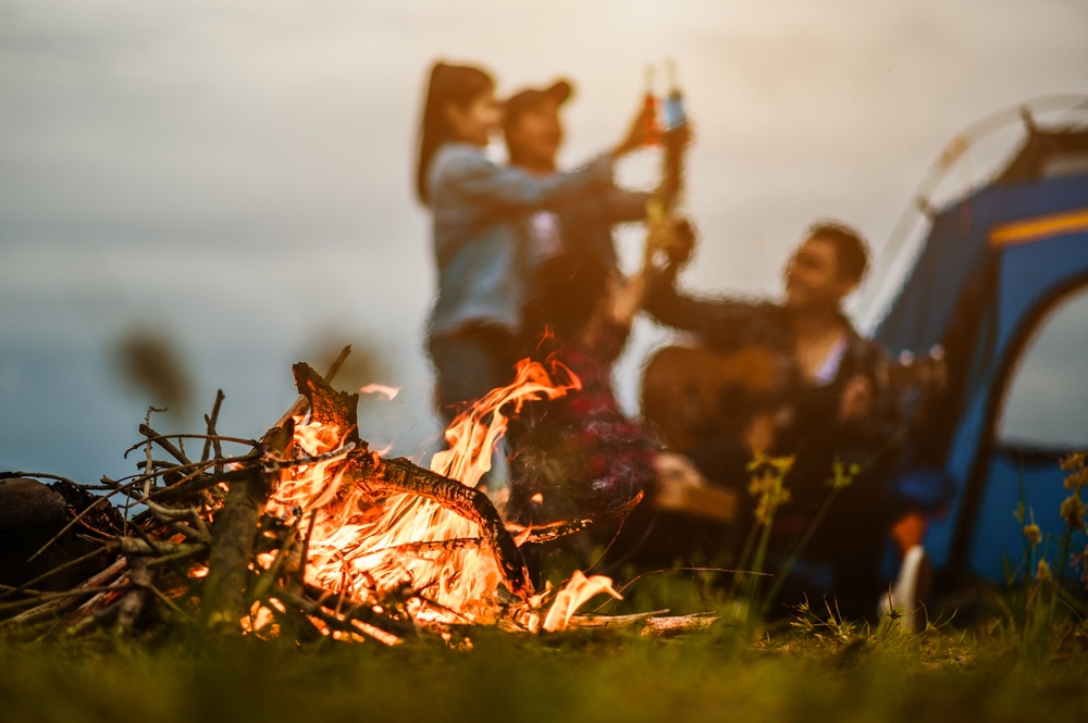 Close-up of a campfire with a tent and group of people in the background.