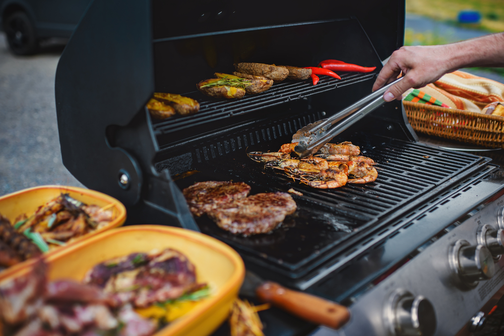 Person using metal tongs to flip various foods on a barbecue.