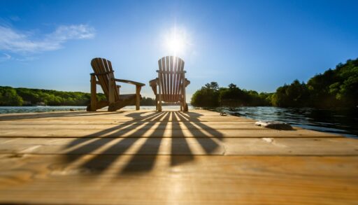 Low angle of sun shining through the back of a Muskoka chair on a dock next to a lake.