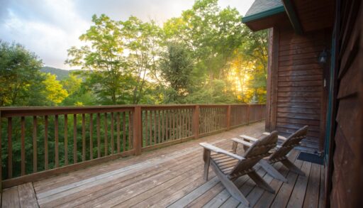 Two Muskoka chairs on a wooden deck overlooking the sun shining through a forest.