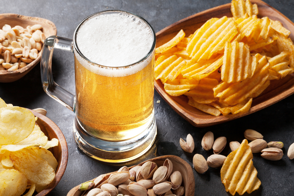 Glass of yellow-coloured beer on a table with chips and peanuts.