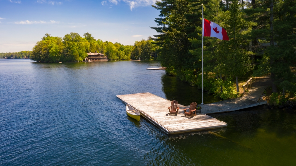 Two Muskoka chairs on a dock with a Canadian flag standing next to it.