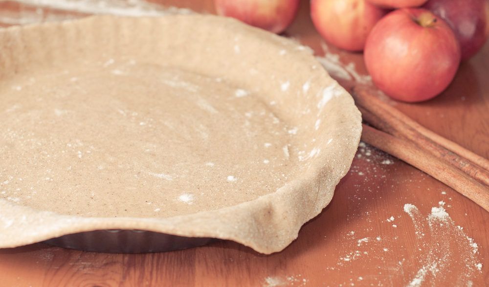 Close-up of an empty pie crust shell with cinnamon sticks and apples on the table next to it.