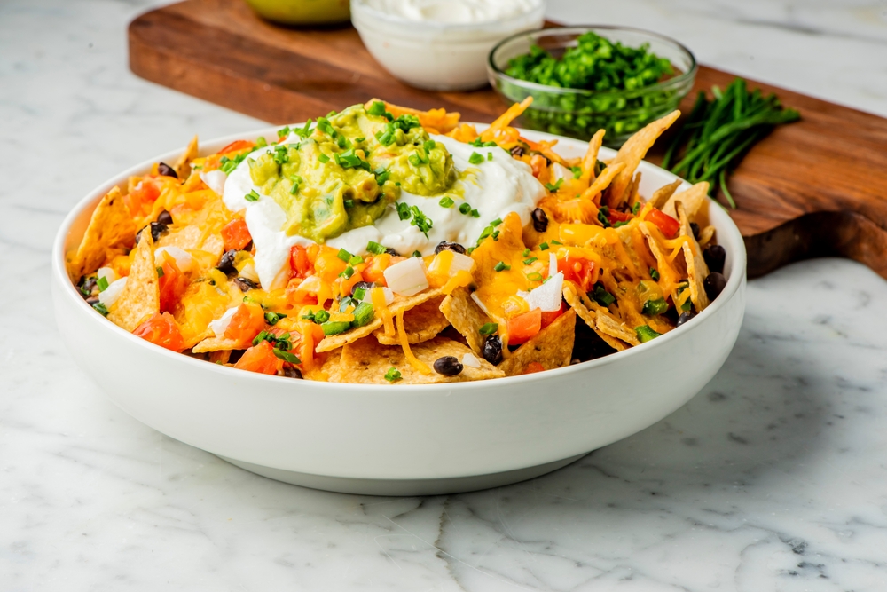 Bowl of nachos loaded with different toppings, next to a wooden cutting board with more toppings.