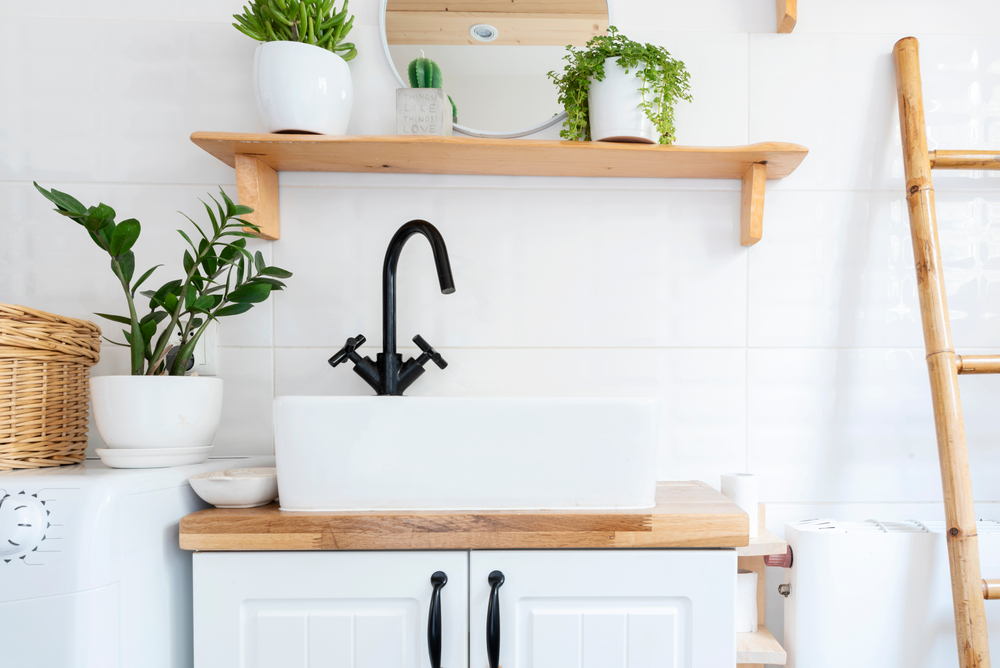 Small, white bathroom with a farmhouse sink, wooden shelving and plants.