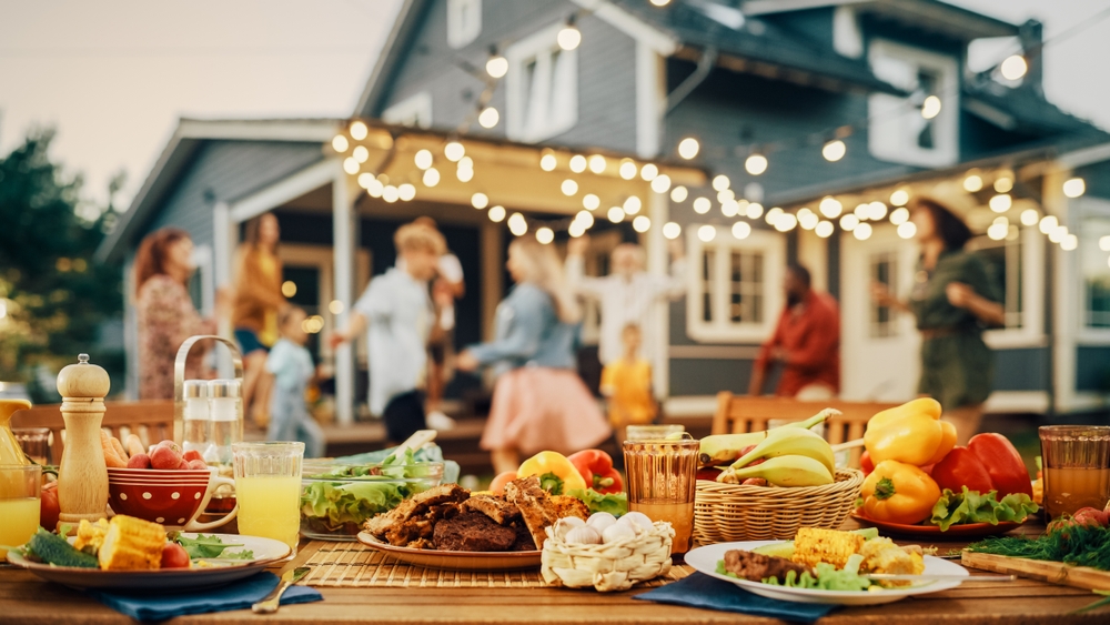 Close-up of an outdoor table with food with a house and group of people in the background.