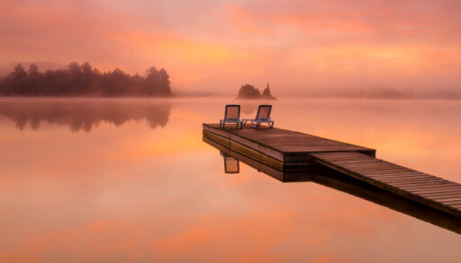 Chairs on a dock with a golden, pink sunrise over a foggy lake.