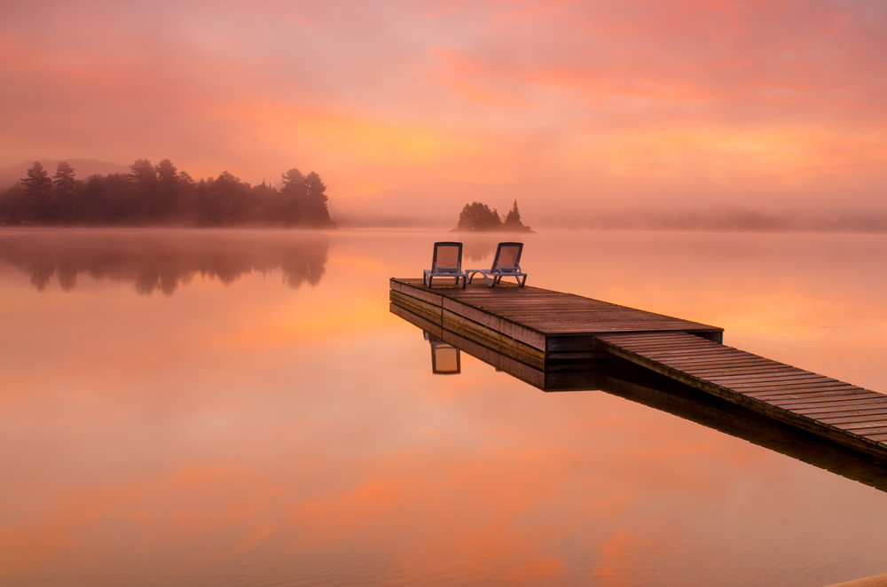 Chairs on a dock with a golden, pink sunrise over a foggy lake.