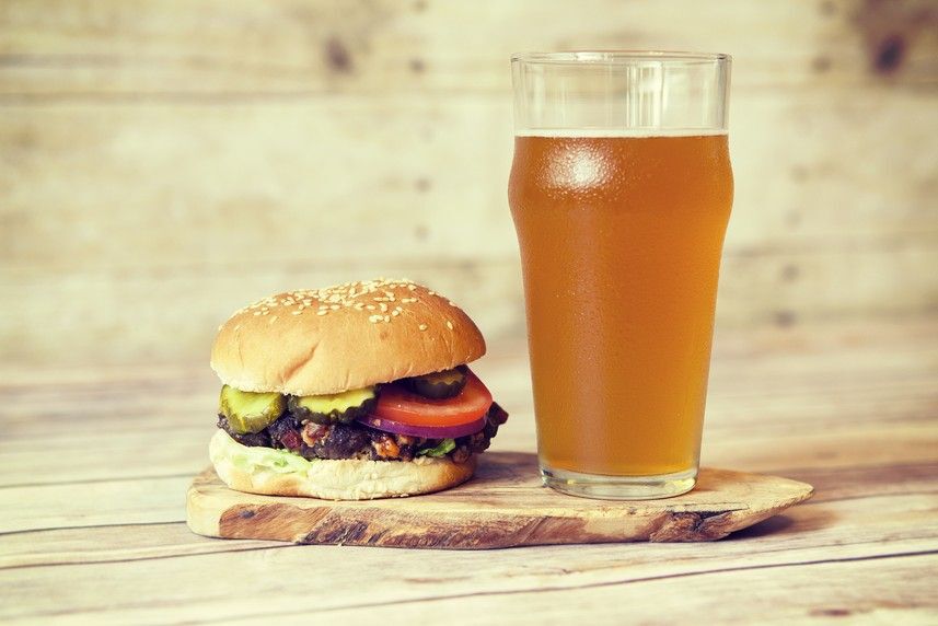 Glass of amber-coloured beer next to a hamburger topped with pickles and tomato.