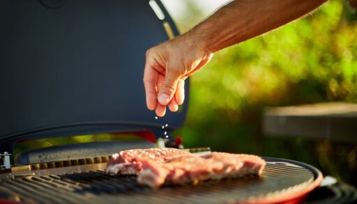 A person sprinkling seasoning over meat on a grill.