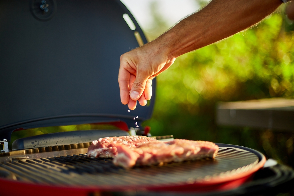 A person sprinkling seasoning over meat on a grill.