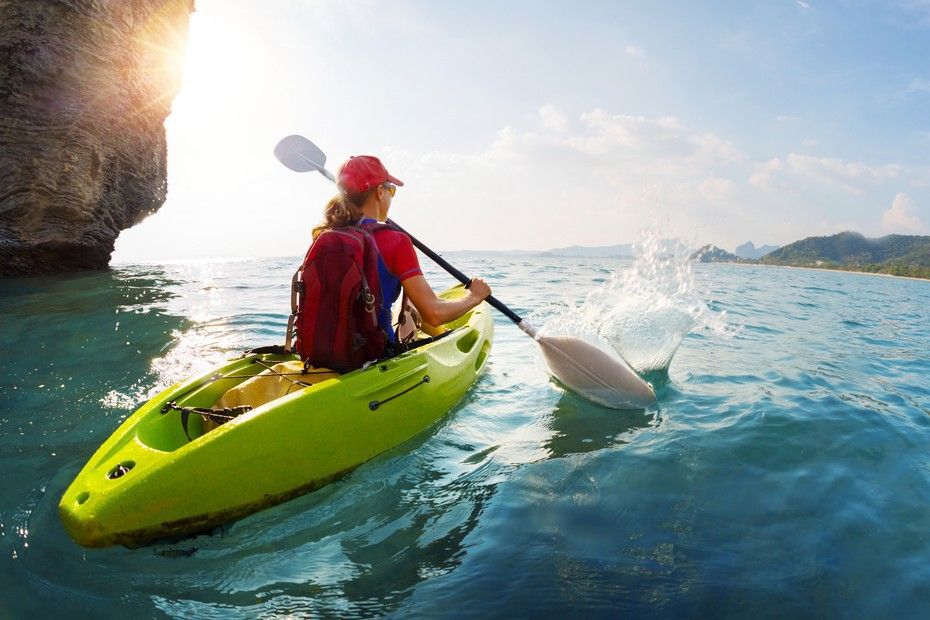 Woman in a red lifejacket and hat paddling a green kayak.