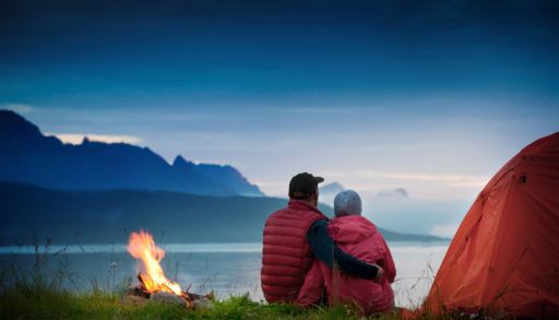 Man and woman sitting next to a tent and campfire looking over a lake surrounded by mountains in the evening.