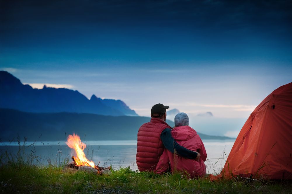 Man and woman sitting next to a tent and campfire looking over a lake surrounded by mountains in the evening.