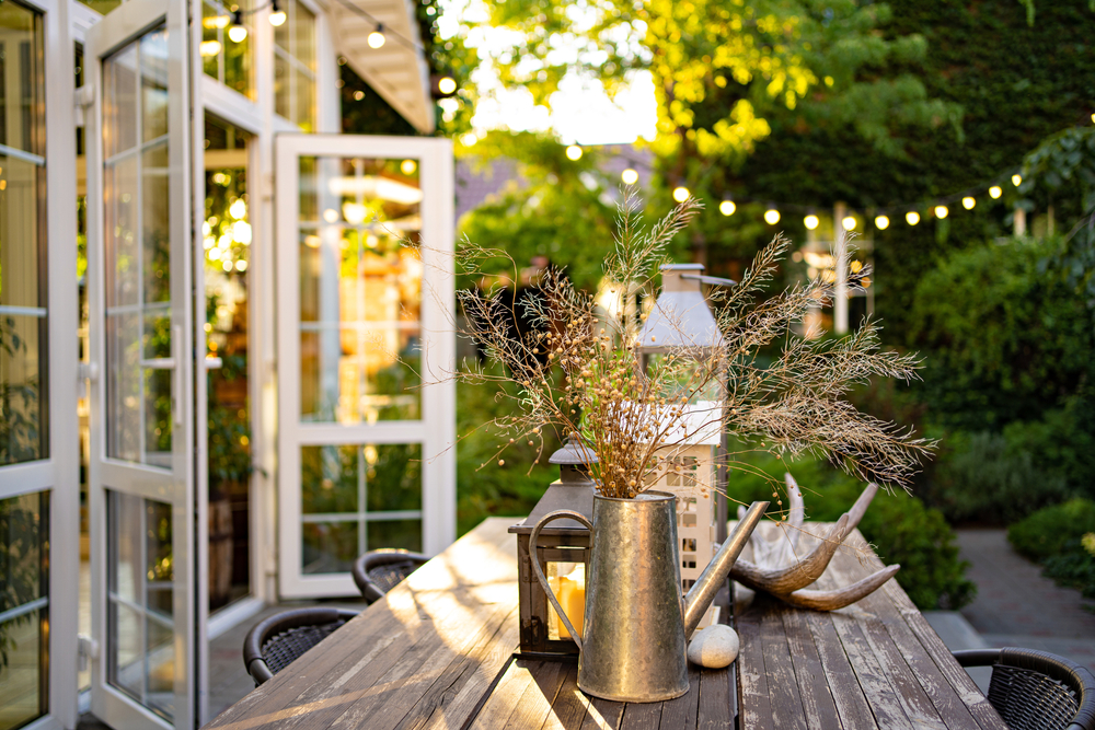 Outdoor dining table with a lantern and metal watering can in the centre and lights hanging overhead.