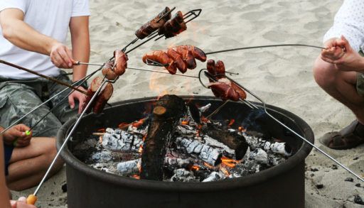 Group of people cooking sausages over a campfire on the beach.